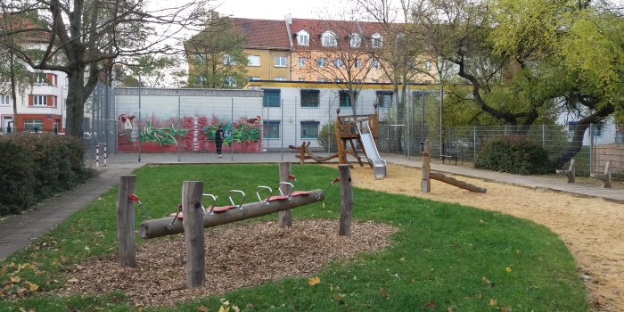 kleiner eingezäunter Bolzplatz, Sandfläche mit Rutschenturm aus Holz und Schaukel mit vier Sitzen