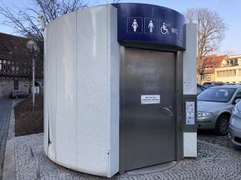 Automatiktoilette mit elliptischen Grundriss, weiß-marmorierter Metallfassade und Edelstahltür  