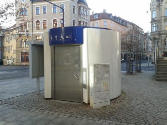 Automatiktoilette mit elliptischen Grundriss, weiß-marmorierter Metallfassade und Edelstahltür  