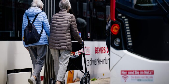 zwei ältere Frauen, davon eine mit Rollator, gehen an einer Haltestelle zwischen zwei Straßenbahnen 