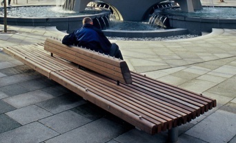 Ein Mann sitzt auf einer braunen Holzbank vor einem Brunnen.