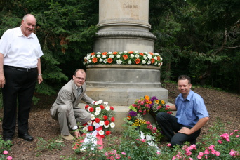 Die Denkmalsäule steht vor eine grünen grünen Hecke und ist mit einem Blumenkranz geschmückt, Der Beigeordnete für Bau und Verkehr Ingo Mlejbnek und der Geschäftsführer des Kakteenzüchtungsunternehmens Ulrich Haage legen Kränze vor der Säule ab. Links neben der Säule steht Dr. Eberhard Czekalla.