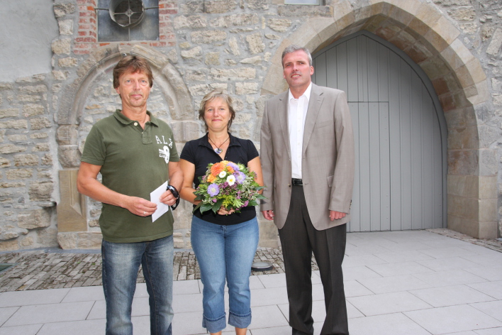 Foto: Oberbürgermeister Andreas Bausewein mit dem Besucherpaar © Stadtverwaltung Erfurt Neben dem Oberbürgermeister Andreas Bausewein stehen links eine Frau und ihr Ehemann. Die Frau hält einen Strauß bunter Blumen in der Hand, der Mann eine Karte. Im Hintergrund ist der Eingangsbereich der Alten Synagoge zu sehen.