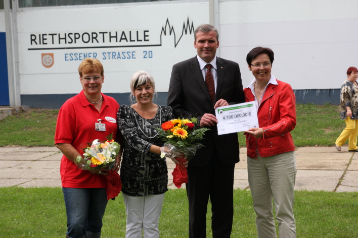 Objektleiterin Rommy Stade, die Vorsitzende des Stadtsportbundes Birgit Pelke, Oberbürgermeister Andreas Bausewein und Sozialministerin Heike Taubert (v.l.n.r.) stehen vor der Riethsporthalle. Der Oberbürgermeister und die Soziaklministerin halten den Fördermittelscheck von 4.100.000,00 Euro vor sich.