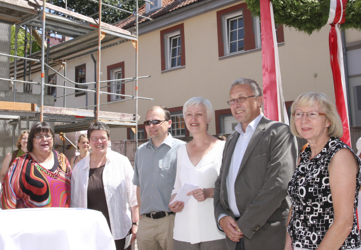 Vor dem Collegium maius steht eine Gruppe von fünf Gästen, links ein Baugerüst und rechts ist ein Stück vom Richtkranz zu sehen.