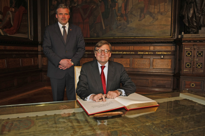 Der dänische Botschafter am Glastisch sitzend bei der Unterschrift im Goldenen Buch, hinter ihm stehend der Oberbürgermeister.