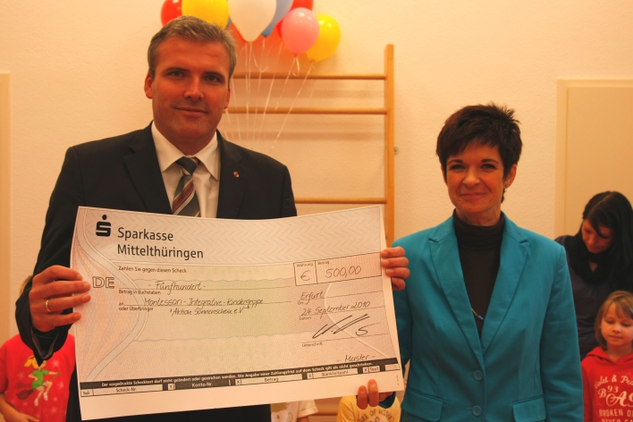 Der Oberbürgermeister Andreas Bausewein hält einen Scheck der Sparkasse Mittelthüringen vor sich. Rechts neben ihm steht die Leiterin der Kinderkrippe Bettina Pokorny. 