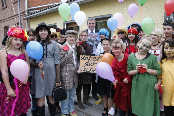 Foto: Die Wiedereröffnung des Jugendzentrums Stotternheim © Stadtverwaltung Erfurt Verkleidete Schülerinnen und Schüler einer Theatergruppe stehen zusammen mit ihrer Lehrerin und dem Oberbürgermeister in einer Gruppe. Der Oberbürgermeister hält im Hintergrund das Einweihungsgeschenk Erfurt-Monopoly in der Hand. Die anderen halten bunte Luftballons hoch. Vorne in der Mitte halten zwei Theaterspieler ein Schild, welches den Titel des Theaterstücks "Unter der Lupe" trägt.