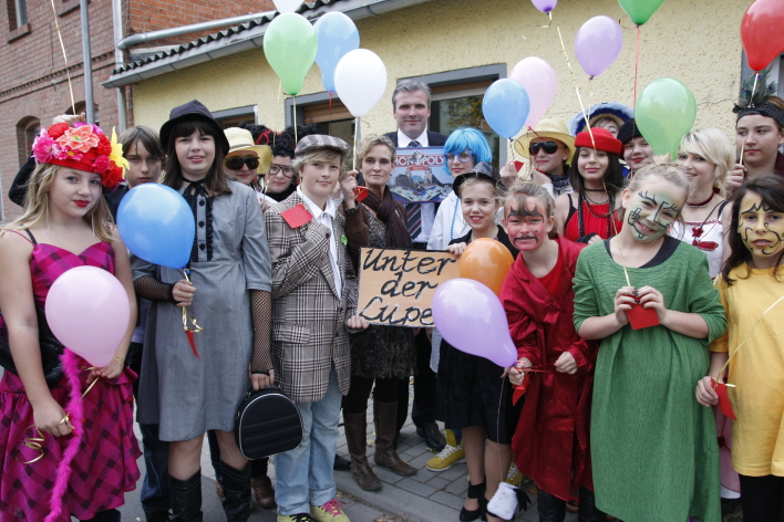 Verkleidete Schülerinnen und Schüler einer Theatergruppe stehen zusammen mit ihrer Lehrerin und dem Oberbürgermeister in einer Gruppe. Der Oberbürgermeister hält im Hintergrund das Einweihungsgeschenk Erfurt-Monopoly in der Hand. Die anderen halten bunte Luftballons hoch. Vorne in der Mitte halten zwei Theaterspieler ein Schild, welches den Titel des Theaterstücks "Unter der Lupe" trägt.