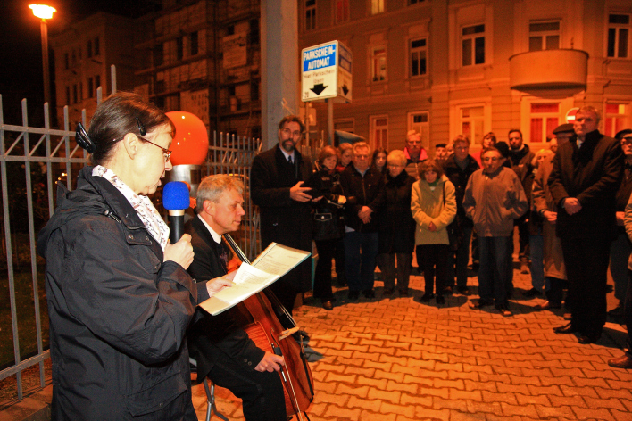 Eine Rednerin, ein Musiker mit Cello und Gäste stehen neben der ca. 1,50 Meter hohen Denknadel auf dem Gehweg.