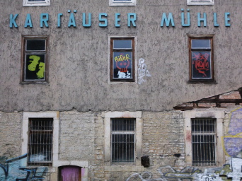 Graue Hausfassade mit bunt bemalten Fenstern unter der Aufschrift "Kartäuser Mühle"