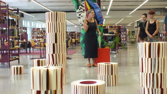 Mehrere Menschen betrachten eine Kunst-Installation mit mehreren Stehlen, die aus kreisförmig, Buchrücken an Buchrücken angeordneten Büchern bestehen. 