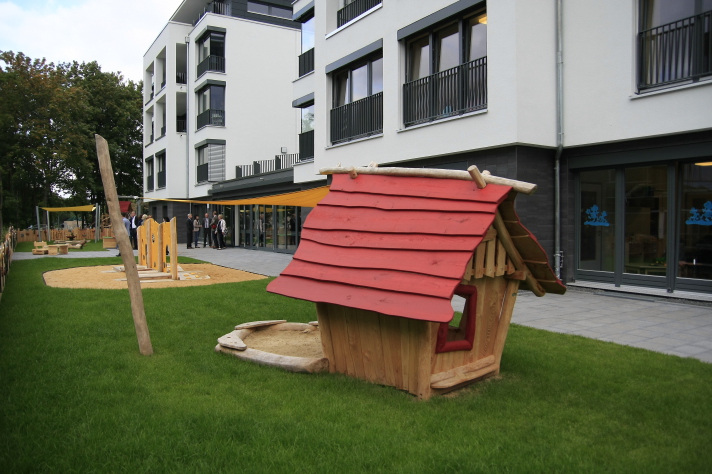 Blick auf das Außengelände. Im Vordergrund ein Spielhaus aus Holz mit Sandkiste und Sonnensegel, im Hintergrund das Kitagebäude. 