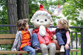 Zwei Kinder sitzen mit der Sandmann-Figur auf einer Bank.
