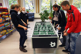 Fotografie: In der Mitte des Bildes steht ein Kickertisch, darum haben sich die Spieler versammelt und kickern