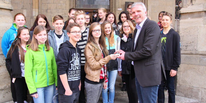 Der Oberbürgermeister übergibt einem jungen Mädchen, die umringt von ihren Mitschülern vor dem Rathaus steht, einen Kristall-Pokal.