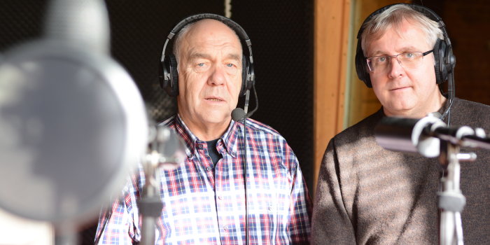Zwei männliche Pesonen mit Kopfhörern hinter Mikrofonen