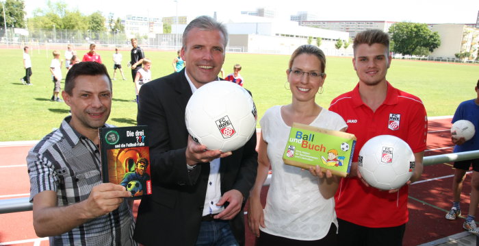 Drei Männer, darunter der Oberbürgermeister, und eine Frau halten zwei Bücher und zwei Fußbälle in die Kamera. Im Hintergrund sieht man Kinder auf einem Sportplatz Fußball spielen. 