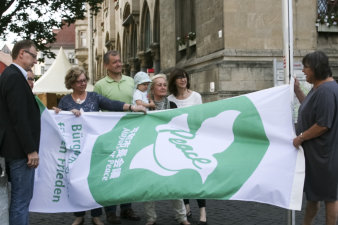 Eine Gruppe aus acht Personen hisst eine grün-weiße Flagge, auf der eine Friedenstaube und die Schriftzüge peace sowie Bürgermeister für den Frieden abgebildet sind. 
