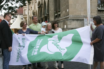 Foto: Bürgermeister für den Frieden © Stadtverwaltung Erfurt Eine Gruppe aus acht Personen hisst eine grün-weiße Flagge, auf der eine Friedenstaube und die Schriftzüge peace sowie Bürgermeister für den Frieden abgebildet sind.
