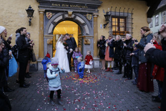 Ein Hochzeitspaar steht vor dem Portal des Hochzeitshauses, um es herum fünf kleine Kinder und die Hochzeitsgesellschaft, die das Paar mit Seifenblasen empfängt. 