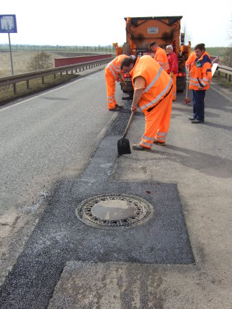 Eine Gruppe männlicher Personen führt auf einer Straße Ausbesserungsarbeiten am Asphalt durch. Sie tragen signalfarbene Arbeitskleidung, im Hintergrund ist ein Transportfahrzeug zu sehen.