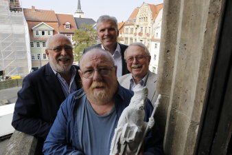 Vier Männer stehen auf einem kleinen Balkon, der vordere Mann hält eine ca. 60 Zentimeter große Figur in der Hand, im Hintergrund sieht man den Fischmarkt. 