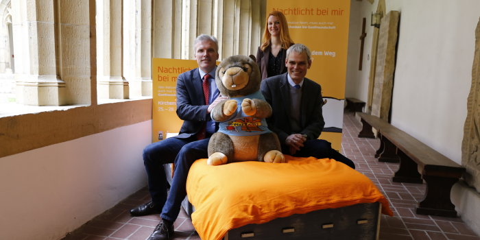 Foto: Kirchtag Bettenkampagne © Stadtverwaltung Erfurt Zwei Männer sitzen in einem Kreuzgang mit einem übergroßen Plüsch-Murmeltier in einem Bett, hinter ihnen steht eine Frau vor einem Werbebanner.