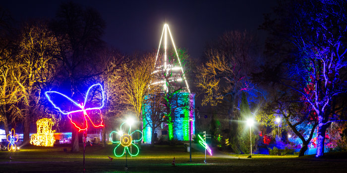 Lichtskulpturen in Form von Schmetterlingen und Blumen leuchten vor illuminierten Bäumen in einem Park. 