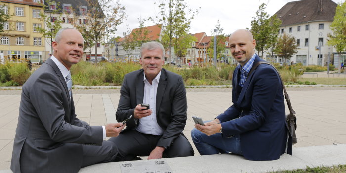 Foto: GPS-Platte Hirschgarten © Stadtverwaltung Erfurt Drei Männer mit Smart-Phones in der Hand sitzen neben einer GPS-Referenzplatte, im Hintergrund sieht man die Parkanlage Hirschgarten.