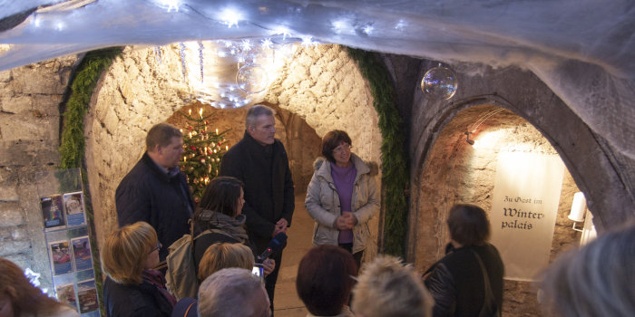 Ein Blick von oben zu einem Gewölbekeller, geschmückt mit Weihnachtsdekoration. Eine Gruppe Menschen, die die Dekorationen anschaut, steht im Fokus.