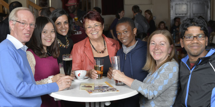 Foto: Internationaler Stammtisch © A. Scholz Viele Personen unterschiedlicher Herkunft stehen lächelnd an einem runden Tisch.