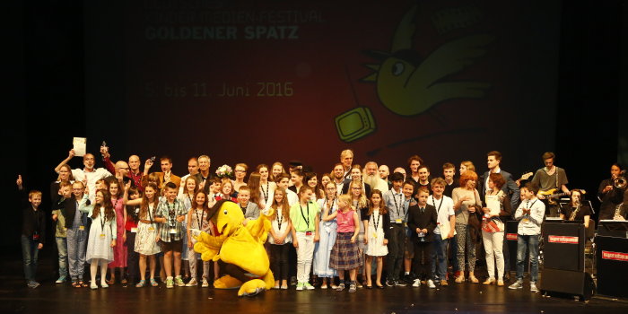 Rund 70 Kinder und Erwachsene stehen auf der Bühne des Theaters Erfurt, einige halten die Trophäe "Goldener Spatz" in den Händen. Rechts von ihnen steht eine Band.