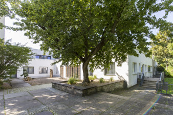 Ein großer Baum steht in der Mitte zwischen einstöckigen, weißen Gebäuden und Sitzmöglichkeiten etwas erhöht und ist das zentrale Motiv des Bildes.