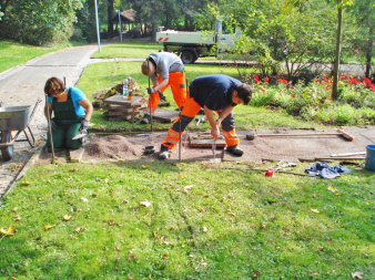 Drei Gärtner in Arbeitskleidung sind dabei einen Weg in einem großen Garten zu erweitern.