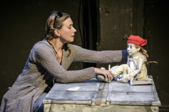 Foto: Theater Waidspeicher © Theater Waidspeicher, Lutz Edelhoff Eine Frau kuzt vor einer Kiste auf der ein weiß gekleidets Handpuppen-Mädchen mit großen Augen und einer roten Strickmütze sitzt.