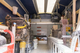 Blick in den mit einer Plane geschlossenen hinteren Teil des Transporters. Dort befinden sich unzählige Arbeitsgeräte und Materialien; ganz hinten steht ein großer Schrank mit Schubfächern, links und rechts stehen beispielsweise ein Notstromaggregat, ein Wasserkanister oder ein Bodenverdichter.