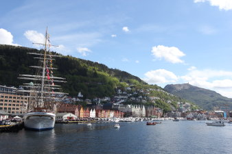 Blick auf die Häuser und das Hafenbecken der norwegischen Stadt Bergen. Links im Bild ist eine große Segelyacht, im Hintergrund sieht man das Hanseviertel Bryggen, die markanten bunten Holzhäuser der Stadt, und den Berg Floyen. 