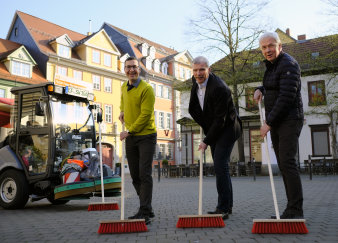 Drei Männer, in der Mitte der Oberbürgermeister, stehen neben einer Kehrmaschine und fegen mit Besen den Platz. 