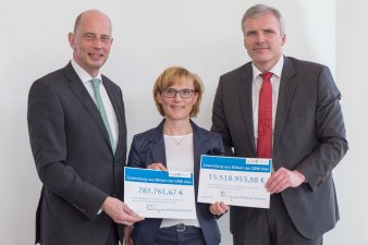 Foto: Danakil Fördermittelbescheid © Stadtverwaltung Erfurt Zwischen zwei Männern steht eine Frau vor einer Leinwand, gemeinsam halten sie zwei symbolische Zuwendungsbescheide in die Kamera.
