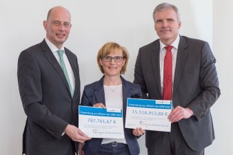 Zwischen zwei Männern steht eine Frau vor einer Leinwand, gemeinsam halten sie zwei symbolische Zuwendungsbescheide in die Kamera. 