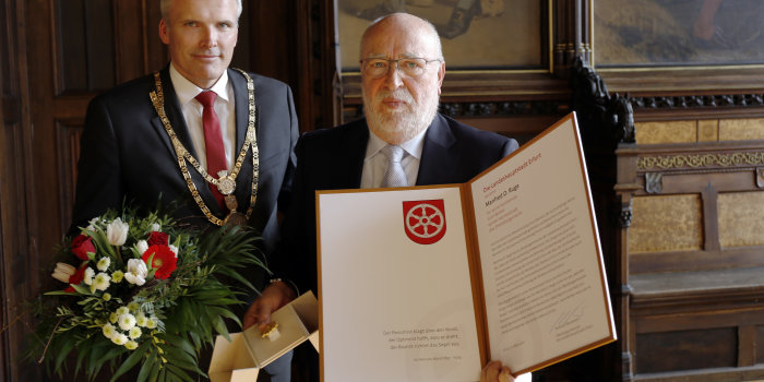 Ein Mann mit Amtskette um den Hals übergibt einem anderen Mann Blumen, ein Geschenk und eine Urkunde.