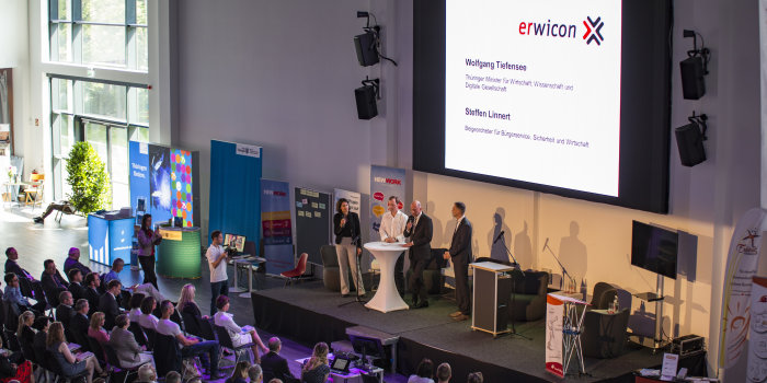 Viele Personen sitzen in einem Saal vor einer Bühne, auf der Bühen sprechen Leute