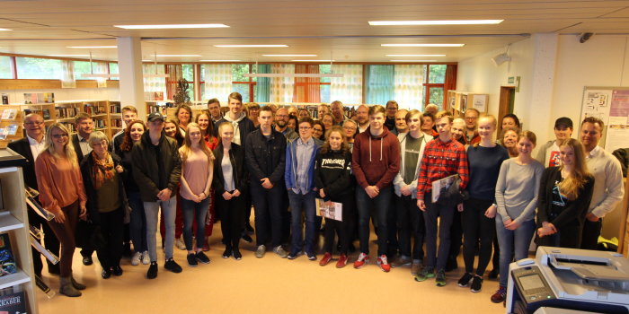 Rund 50 Personen - Schüler und Erwachsene - stehen im Halbkreis für ein Gruppenfoto in einer Bibliothek. 