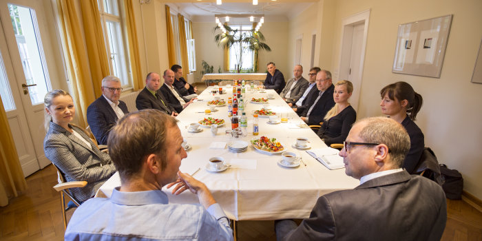 Viele Menschen sitzen an einem Tisch und hören einem Redner zu.  