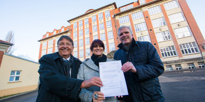 Foto: Übergabe des Fördermittelbescheides © Bild13 Zwei Herren und eine Damen stehen vor einem Schulgebäude, alle gemeinsam halten ein Dokument in ihren Händen.
