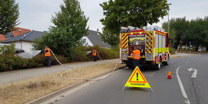 Drei Feuerwehrmänner mit Löschschlauch und einem Einsatzfahrzeug.