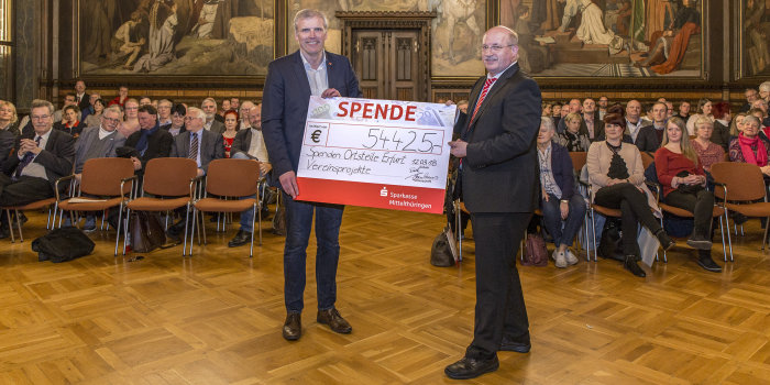 Zwei Männer halten ein symbolischen Scheck in ihren Händen, darauf steht die Summe von 54.425 EUR.