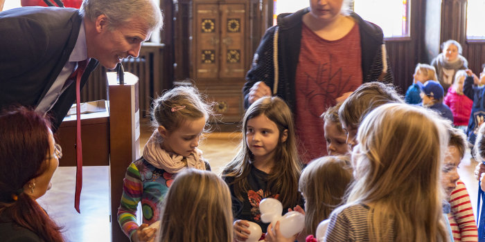 Ein großer Mann beugt sich zu vielen kleinen Kindern, die zum Teil eine weiße Plastikente in ihren Händen halten. 