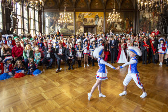 Ein Kinderpaar tanzt vor vielen Leuten mit Narrenkappen in einem historischen Saal.